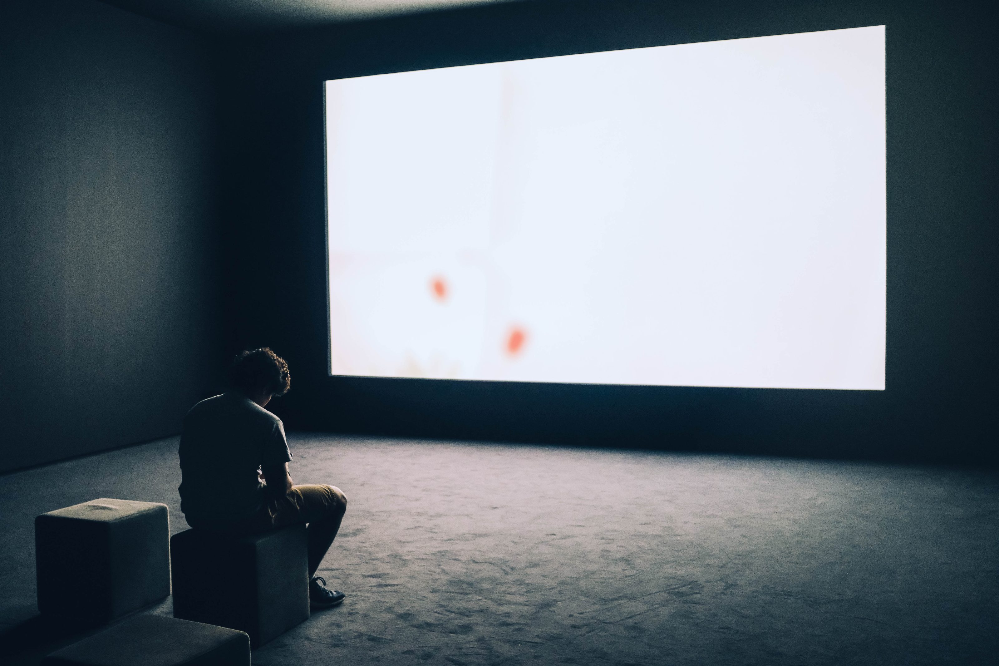 A lone viewer seated in front of a lit projection screen in a darkened room.