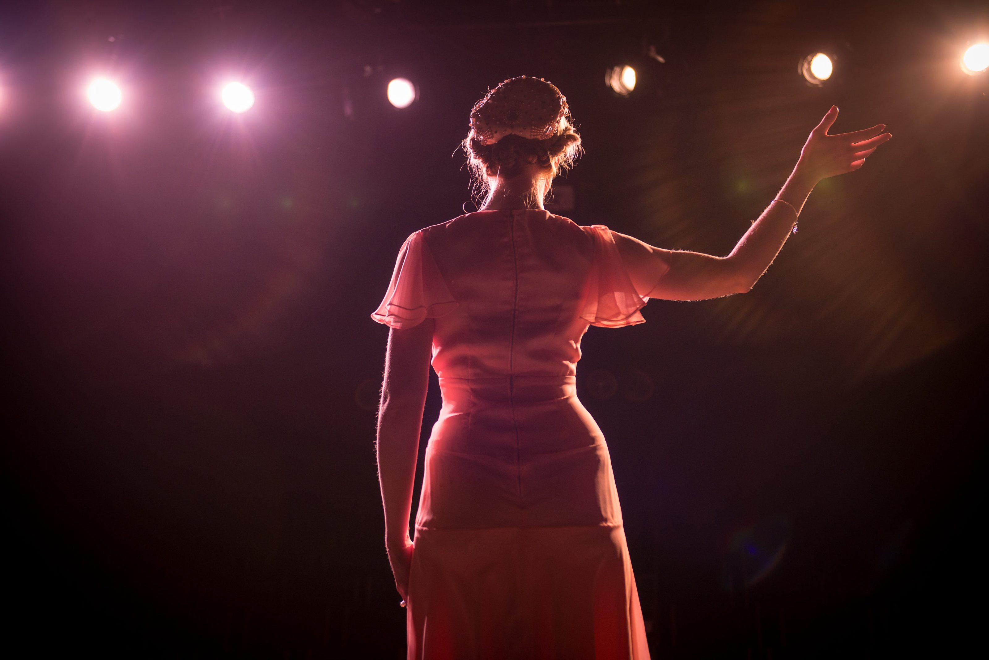 A performer on stage with one arm raised, backlit by theatre lights.
